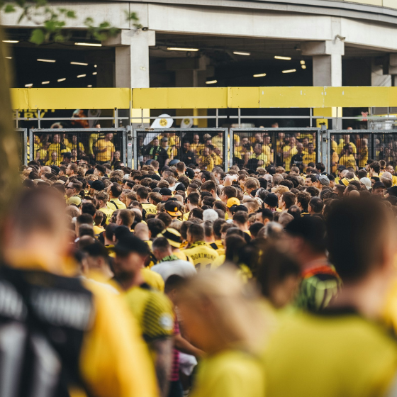 A large crowd of people wearing yellow shirts are gathered outside a stadium entrance with metal gates.