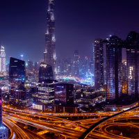 Night skyline of a modern city with a tall central skyscraper, illuminated high-rises, and long-exposure light trails from busy highways curving through the foreground.