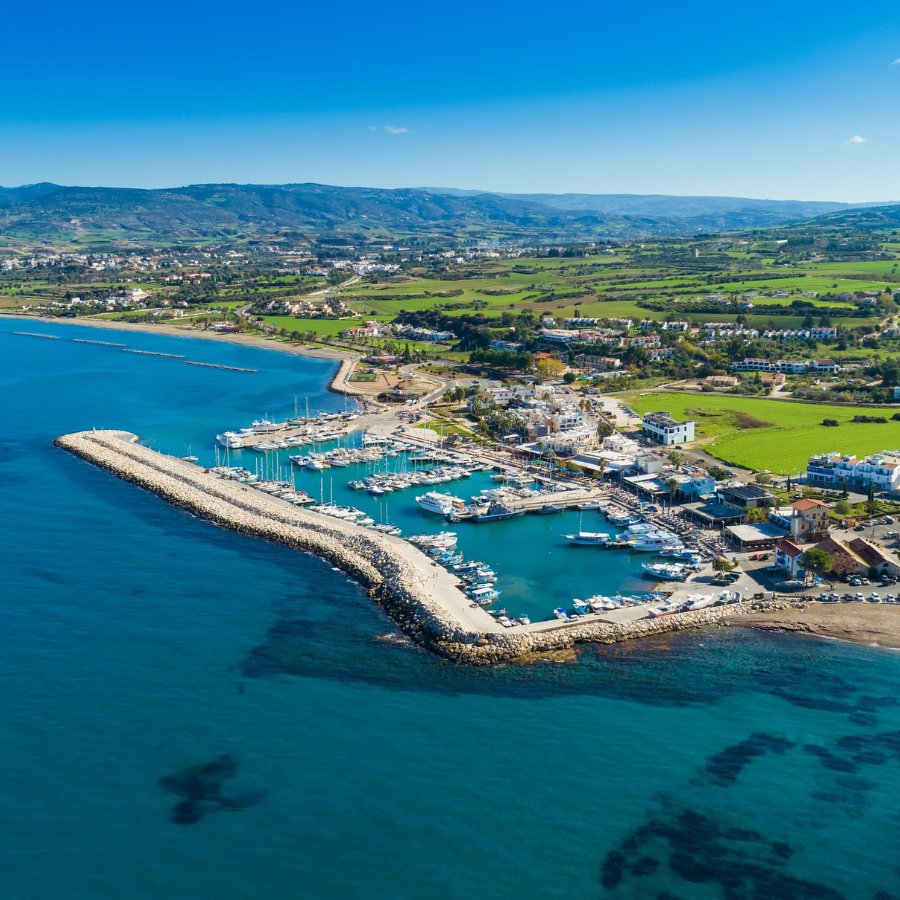Aerial view of Latchi, a Cypriot coastal town with a long breakwater, marina docks, sandy shoreline, and green fields stretching inland under a blue sky.