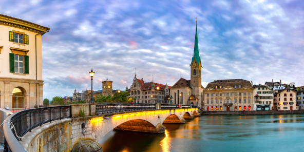 Evening view of a riverside bridge with illuminated arches over calm water, lined with historic buildings and a tall green church spire under a cloudy blue sky.