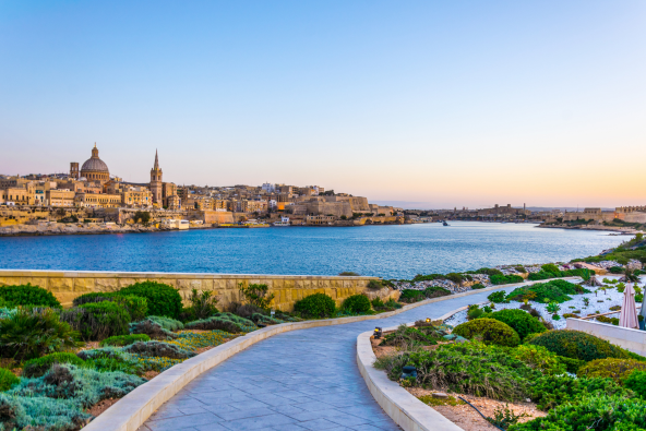 Golden-hour view of a coastal city with historic buildings and domes along the shoreline, a curving seawall, blue water, and a landscaped walkway with shrubs under a pastel sunset sky.