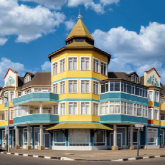 A brightly coloured multi-story building with a central golden tower and arched windows stands on a street corner under a blue sky with scattered clouds.