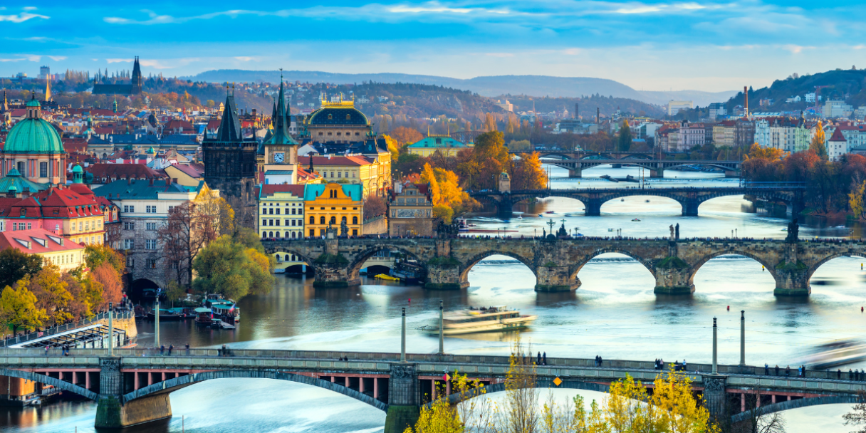 Panoramic view of a riverside city at sunrise with autumn-coloured trees, historic buildings, and multiple arched bridges spanning the wide river.