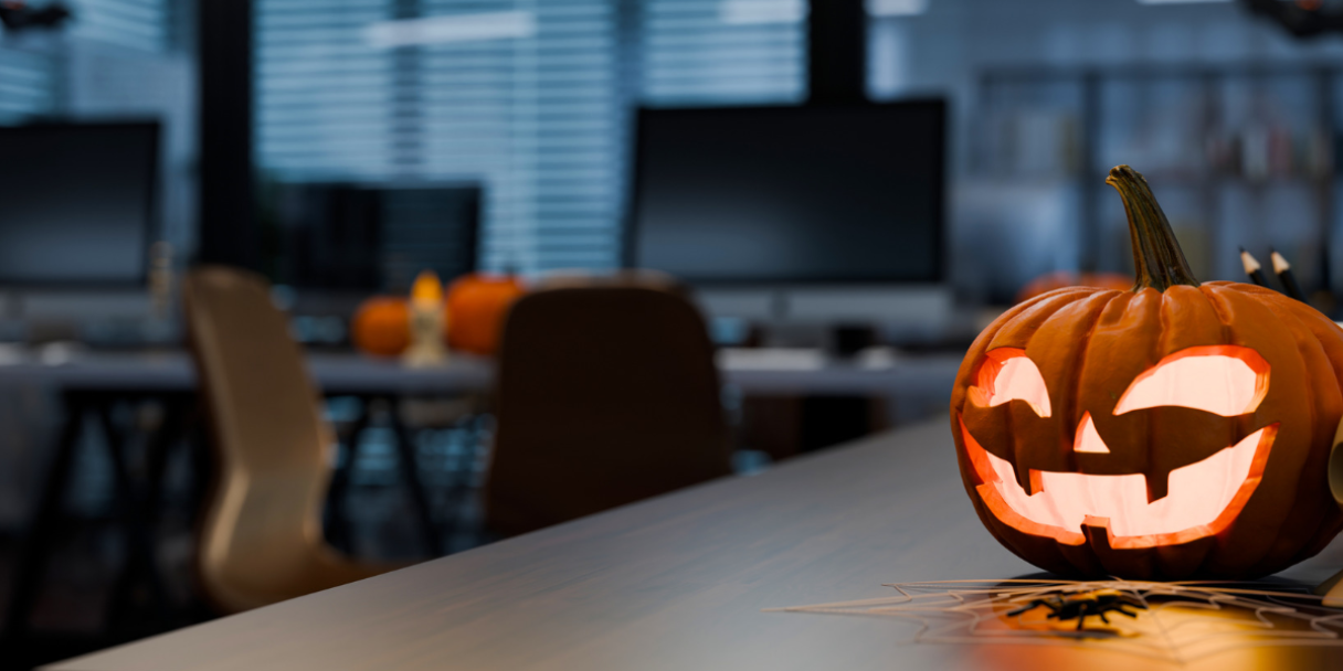 Halloween decorations on a desk: a carved jack-o’-lantern with glowing eyes and mouth, a small skull figure, and a few scattered pumpkin pieces in a softly blurred office with monitors and chairs.