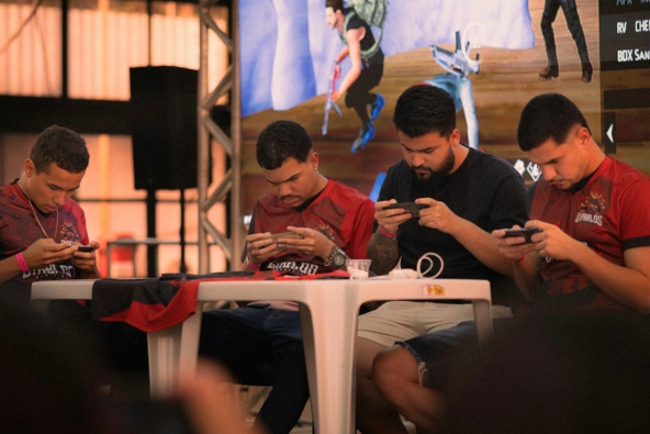 Four individuals are seated at tables, engrossed in playing mobile games. The background features a large screen displaying game graphics.