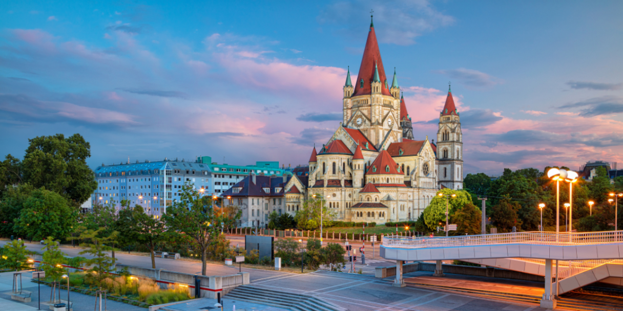 A grand church with red roofs and spires stands behind a modern plaza with a fountain and streetlights at dusk.