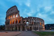 Colosseum in Rome at dusk, illuminated with warm lights, viewed from a stone walkway under a deep blue evening sky with scattered clouds.