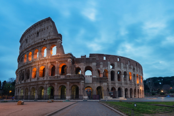 Colosseum in Rome at dusk, illuminated with warm lights, viewed from a stone walkway under a deep blue evening sky with scattered clouds.