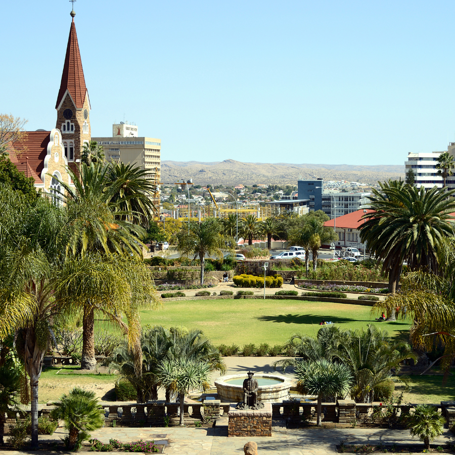View of a landscaped park with palm trees and a stone walkway, overlooking city buildings and a tall church with a red spire under a clear blue sky.