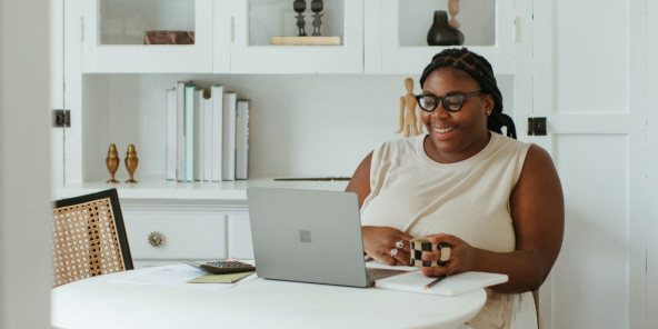 A smiling Black female worker, dressed in a white outfit and holding a coffee mug with both hands, while working from home