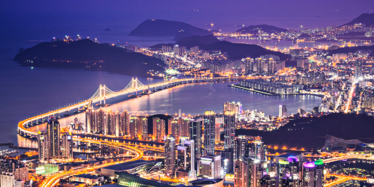 a panoramic night time view of a Busan with illuminated skyscrapers and a large suspension bridge spanning a body of water