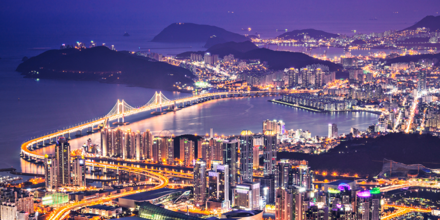 a panoramic night time view of a Busan with illuminated skyscrapers and a large suspension bridge spanning a body of water