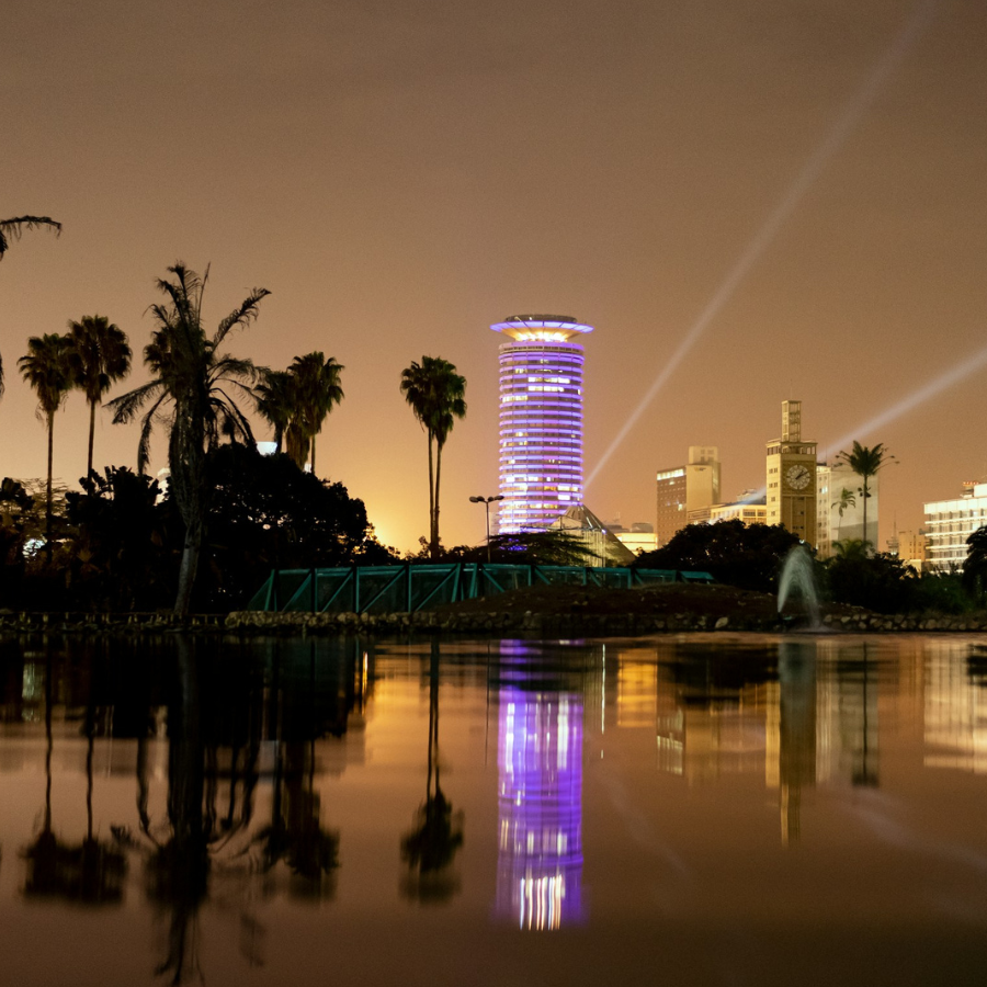 A nighttime cityscape with palm trees in the foreground and illuminated skyscrapers reflected in a body of water.