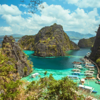 A scenic view of a tropical bay with turquoise water, surrounded by lush green islands and karst cliffs. Numerous small boats are moored in the clear water, and a pathway is visible along the shore.