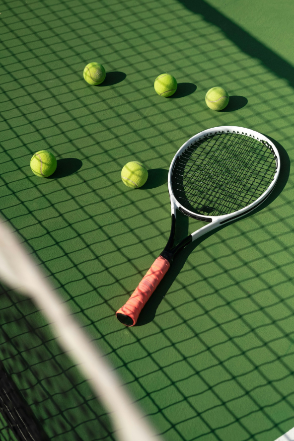 A tennis racket rests on a green court next to six tennis balls, with a net in the foreground casting shadows.