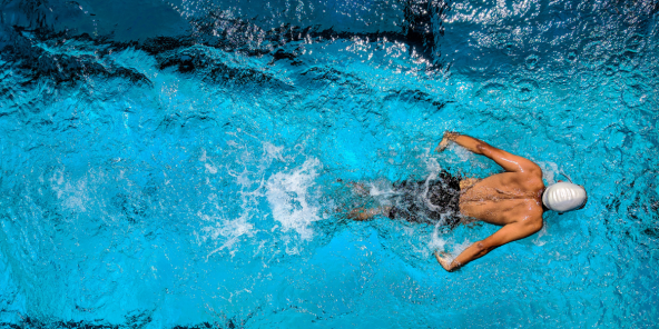 A swimmer in a white cap and black shorts swims the butterfly stroke in bright blue water, creating a splash.