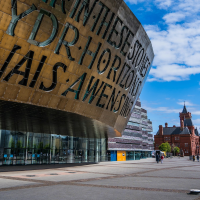 a large, curved building with text inscribed on its surface, set against a bright blue sky with fluffy clouds. In the background, a red brick building and a Ferris wheel are visible