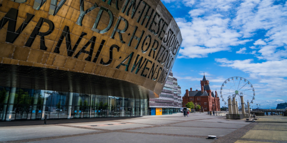 a large, curved building with text inscribed on its surface, set against a bright blue sky with fluffy clouds. In the background, a red brick building and a Ferris wheel are visible