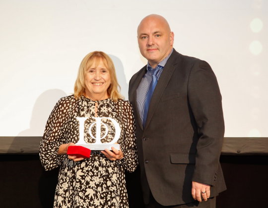 Ann in a black and white patterned dress holds a white trophy with "IoD" inscribed on it, while a man in a suit and blue tie stands beside her.