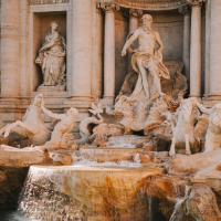 Sunlit view of a grand historic building facade with ornate stone statues and archways above a turquoise fountain pool, with water splashing at the base under a blue sky.