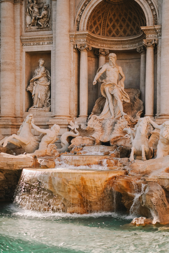 Sunlit view of a grand historic building facade with ornate stone statues and archways above a turquoise fountain pool, with water splashing at the base under a blue sky.