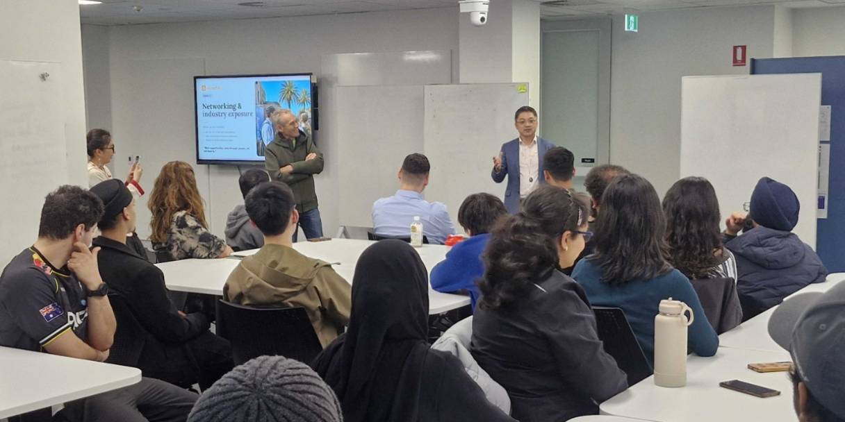 Smiling leaders in the foreground at a conference classroom, with rows of chairs and multiple projector screens showing slide presentations in the background.