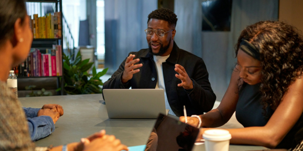 A man speaks and gestures while presenting on a laptop to coworkers seated around a conference table, with notebooks, phones, and a takeaway coffee cup nearby in a modern office.
