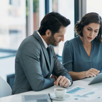a Caucasian man and woman in business attire looking at a tablet computer and documents on a table in an office setting.