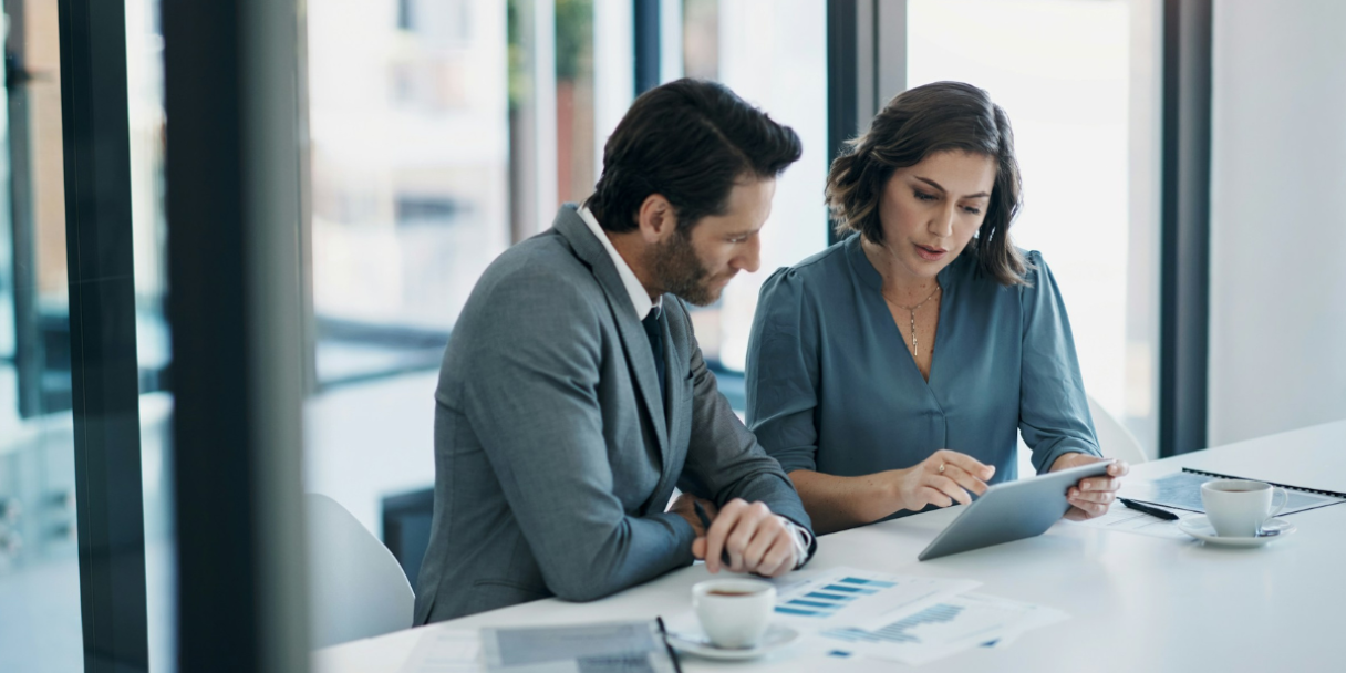 a Caucasian man and woman in business attire looking at a tablet computer and documents on a table in an office setting.
