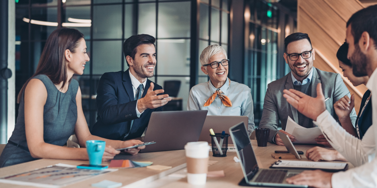 a group of professionals in business attire are gathered around a conference table, engaged in a lively discussion, with laptops and documents spread out before them