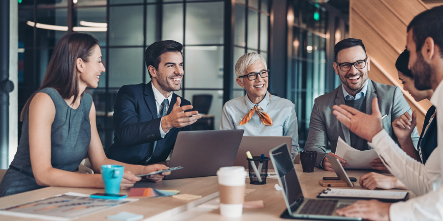 a group of professionals in business attire are gathered around a conference table, engaged in a lively discussion, with laptops and documents spread out before them