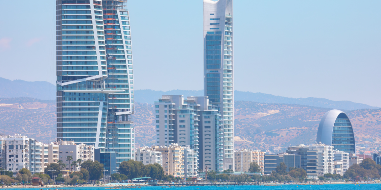 Modern skyscrapers, including one under construction with a crane, line a waterfront with blue water in the foreground and hills in the background.