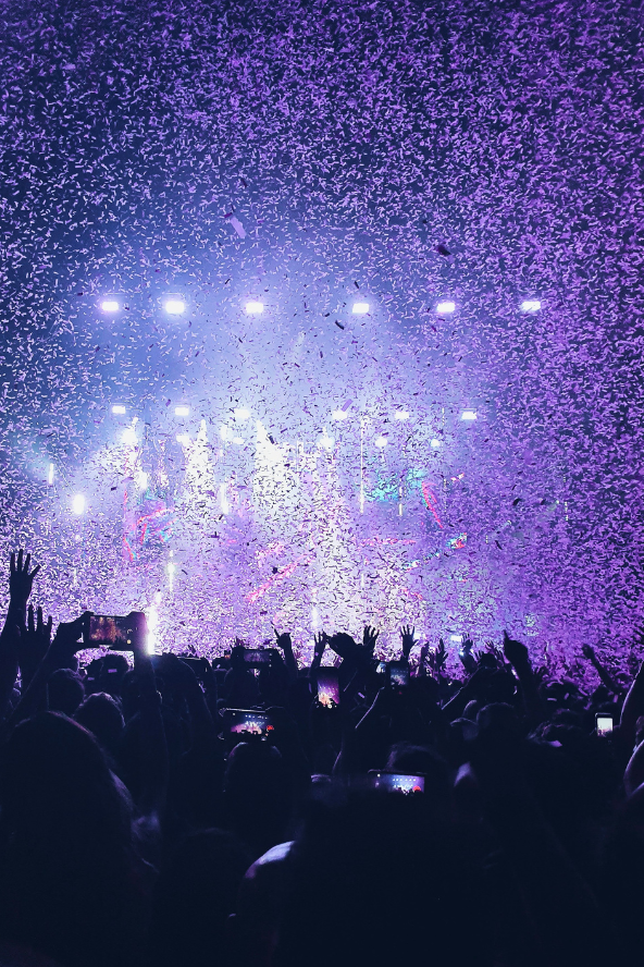 A crowd of people with their hands raised at a concert, with purple confetti falling from above and bright stage lights in the background.