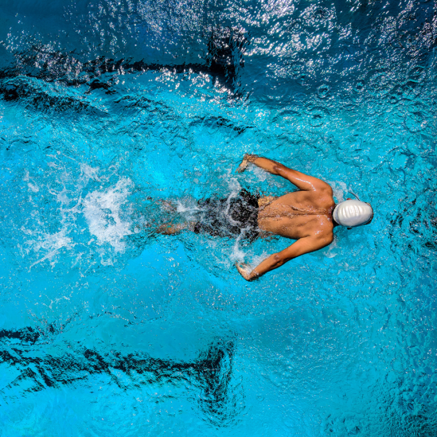 A swimmer in a white cap and black trunks swims butterfly stroke in a bright blue pool.