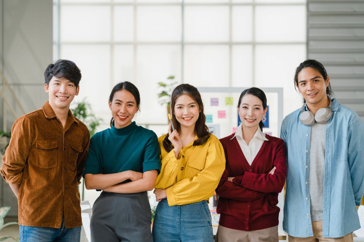 Group of Asia young creative people in smart casual wear smiling and arms crossed in creative office workplace. Diverse Asian male and female stand together at startup. Coworker teamwork concept.