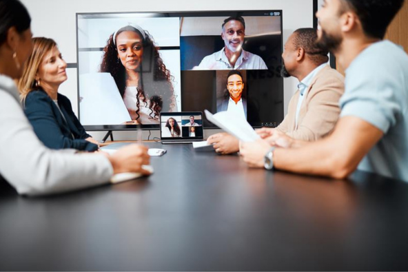 A group of people in a meeting room participate in a video conference call displayed on a large screen and a laptop.