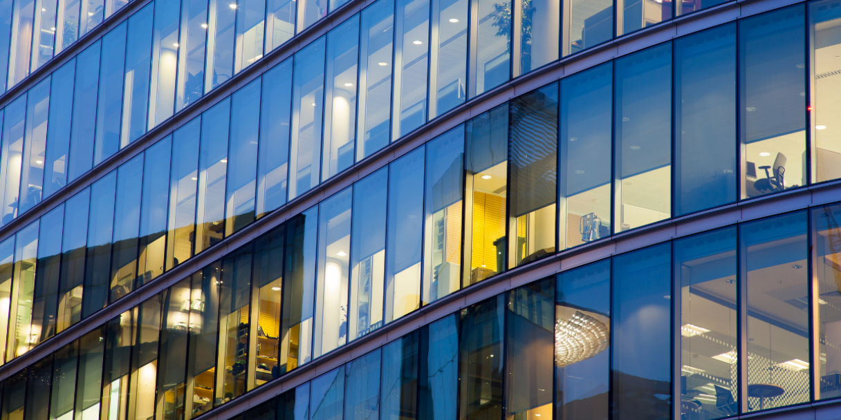 A modern office building with rows of glass windows, some illuminated from within, reflecting the blue evening sky.