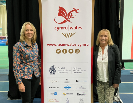 Two women stand next to a large banner that says "Cymru | Wales", along with logos of sponsors. The banner has a red dragon emblem at the top.
