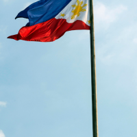 The flag of the Philippines, with its blue and red stripes and white triangle containing a golden sun and stars, waves on a flagpole against a bright blue sky with scattered white clouds.