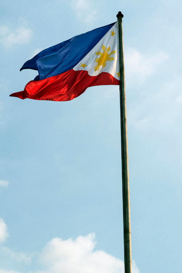 The flag of the Philippines, with its blue and red stripes and white triangle containing a golden sun and stars, waves on a flagpole against a bright blue sky with scattered white clouds.