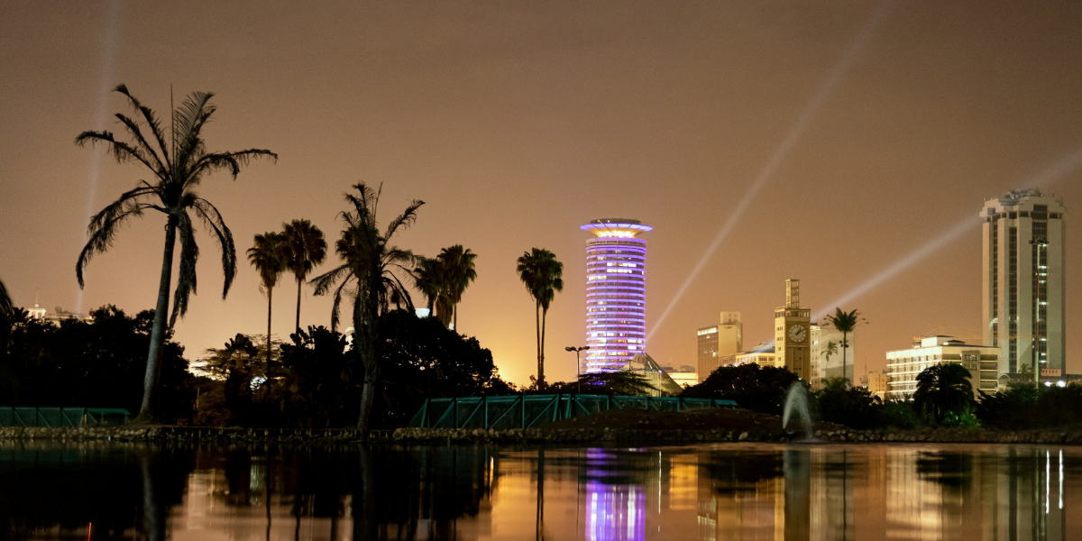 A city skyline at night with tall buildings, palm trees, and a body of water reflecting the lights.
