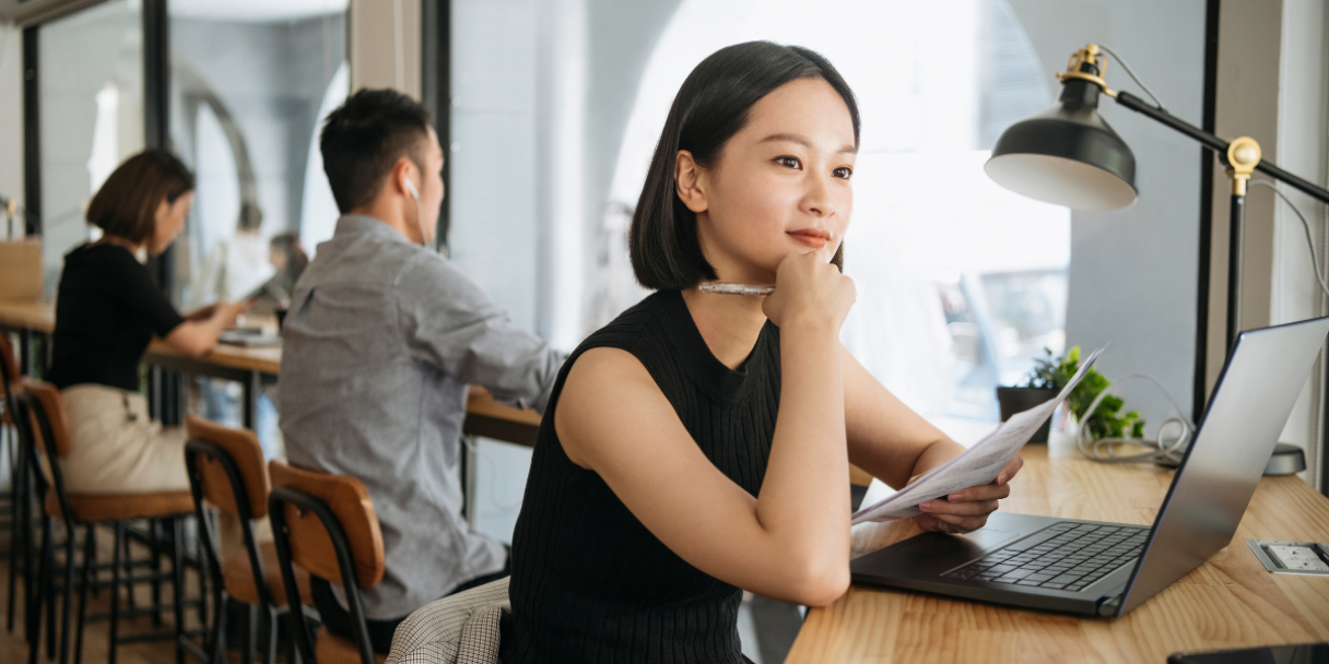 a Chinese woman sits at a desk with a laptop and papers, resting her chin on her hand and looking away from the camera