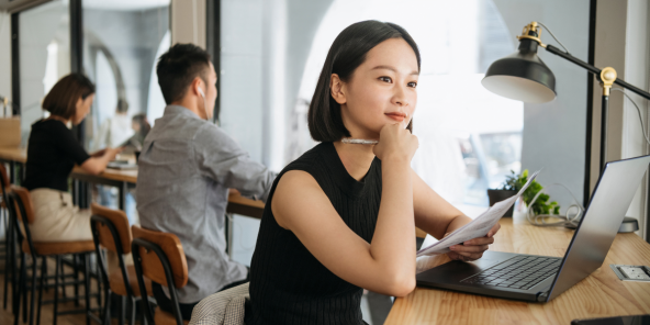 a Chinese woman sits at a desk with a laptop and papers, resting her chin on her hand and looking away from the camera
