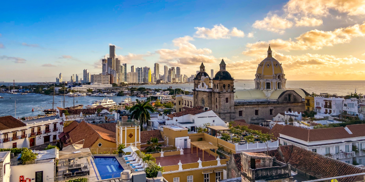 A panoramic view of the historic walled city of Cartagena, Colombia, with its colonial architecture and domes in the foreground and a modern skyscraper skyline in the background under a bright blue sky with scattered clouds.