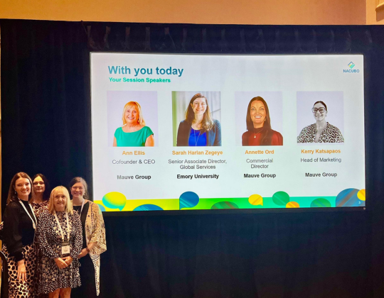 Four women stand in front a digital screen displaying their headshots and job titles.
