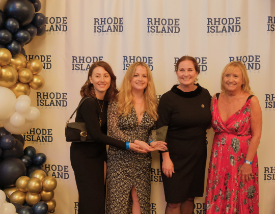 Four women stand in front of a backdrop with "Rhode Island Commerce" repeated, posing for a photo with balloons to the left and right.
