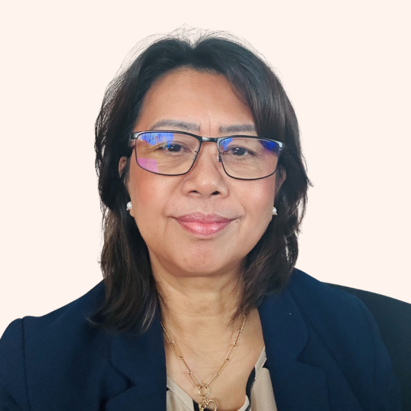 Portrait of a person with shoulder-length dark hair wearing blue-framed glasses, a dark blazer, and a layered gold necklace, facing the camera against a pale yellow background.