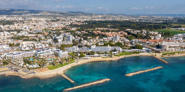 Aerial view of Paphos with dense white buildings along a sandy shoreline, clear turquoise water, and a small breakwater extending into the sea.