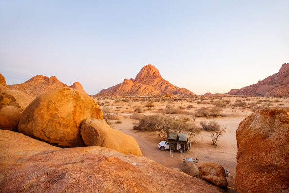 Two camping chairs and a small table sit in a desert valley between large orange rock formations, with sparse shrubs and distant mountains under a pale sky at sunrise.