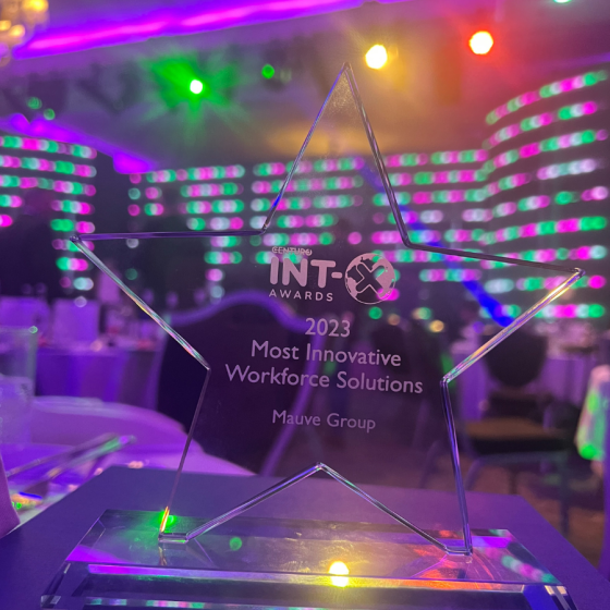 a star-shaped glass award engraved with '2023 Most Innovative Workforce Solutions, Mauve Group' sits on a table with a blurred background of a banquet hall with colourful lights